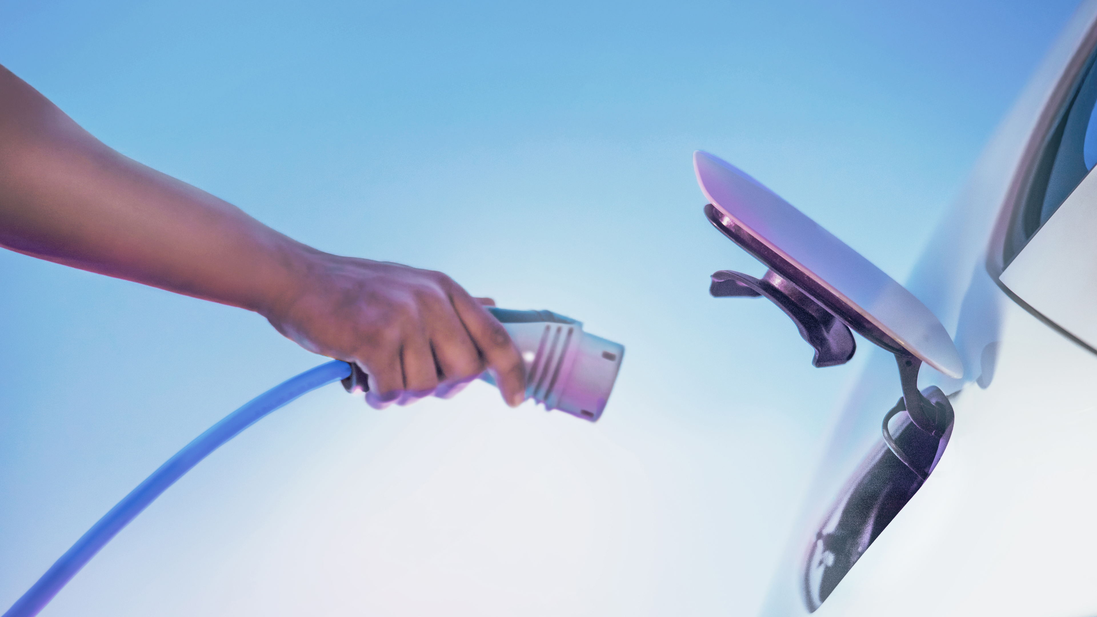 Ein Mensch verbindet einen Ladestecker mit einem Elektroauto unter klarem, blauem Himmel – Symbol für nachhaltige Mobilität.